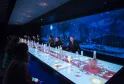 Sala de restaurante futurista con mesa interactiva iluminada, vista panorámica nocturna con luna y árboles en pantalla gigante, comensales sentados a ambos lados
