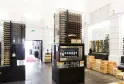 Interior moderno de la galería Max Bordeaux Wine Gallery & Cellar con vitrinas iluminadas de botellas de vino, cajas de madera apiladas y equipos de degustación en un espacio luminoso y minimalista.