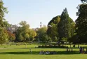 Vista del Jardín Público de Burdeos con césped verde, árboles frondosos, visitantes descansando, un monumento con estatua al fondo y edificios históricos bajo cielo despejado.