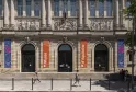 Fachada del Museo de Aquitania en Burdeos con columnas imponentes, relieves esculpidos, pancartas naranjas y azules, tres puertas arqueadas y visitantes en la plaza.